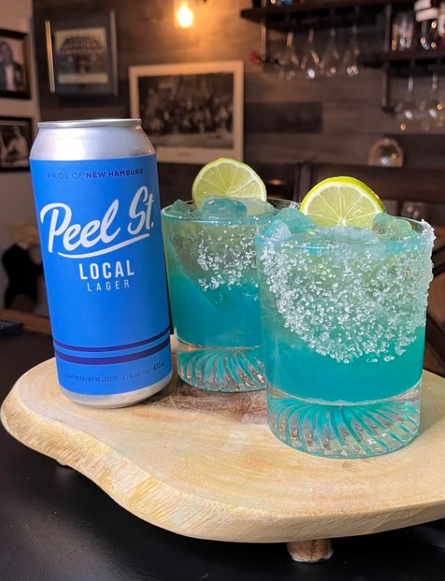 Salt-rimmed tumbler glass half-filled with vibrant blue tequila-lime mix over ice, topped with foamy Peel St Lager; lime wheel garnish perched on the edge, evoking tropical beach vibes

