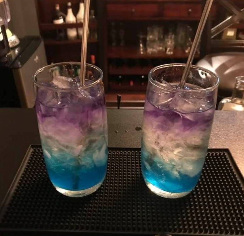 Hand gently floating purple Empress Gin over white wine in an ice-topped wine glass with blue curacao base; Cointreau bottle and optional lemon juice nearby, bubbles subtly rising for a mesmerizing pour.

