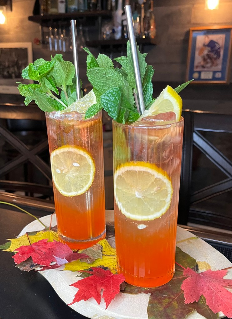 A vibrant orange and red layered cocktail in a glass, named "The Autumn Colours Cocktail" The drink features Empress Gin, Aperol, and honey, evoking autumn vibes with bursting flavors.