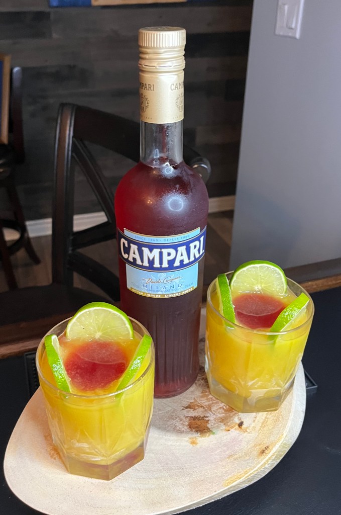 A tumbler full of a stunning red and orange cocktail with the red in the center of the glass.  Made with frozen OJ and lime with Campari and Sprite - handcrafted, vibrant and very tasty!