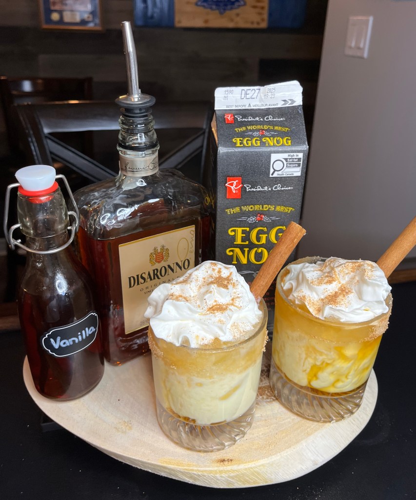 Two tumblers filled with a hand-crafted cocktail of Disaronno, Vanilla, Egg Nog with a whip cream topping and cinnamon stick sit on a rustic wooden tray.
