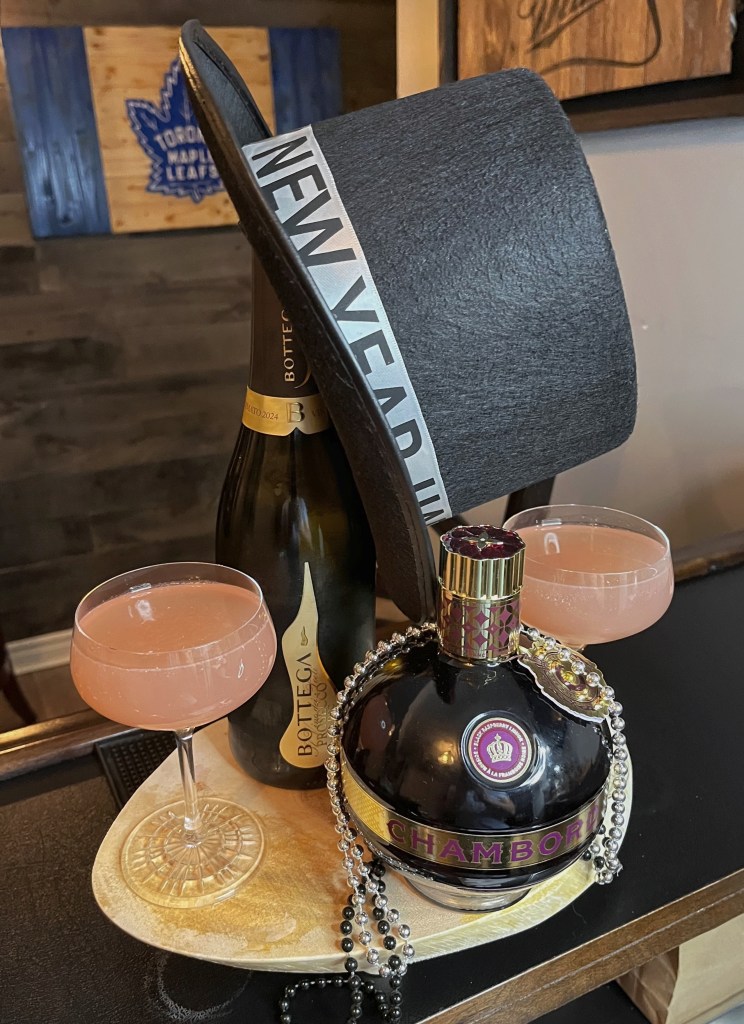 This hand crafted New Years Eve Cocktail sits on a wooden board and is pale pink in color. A large New Year's top hat hangs on top of a bottle of sparkling wine and a bottle of Chambord. The image gives off a New Year's celebration feel.