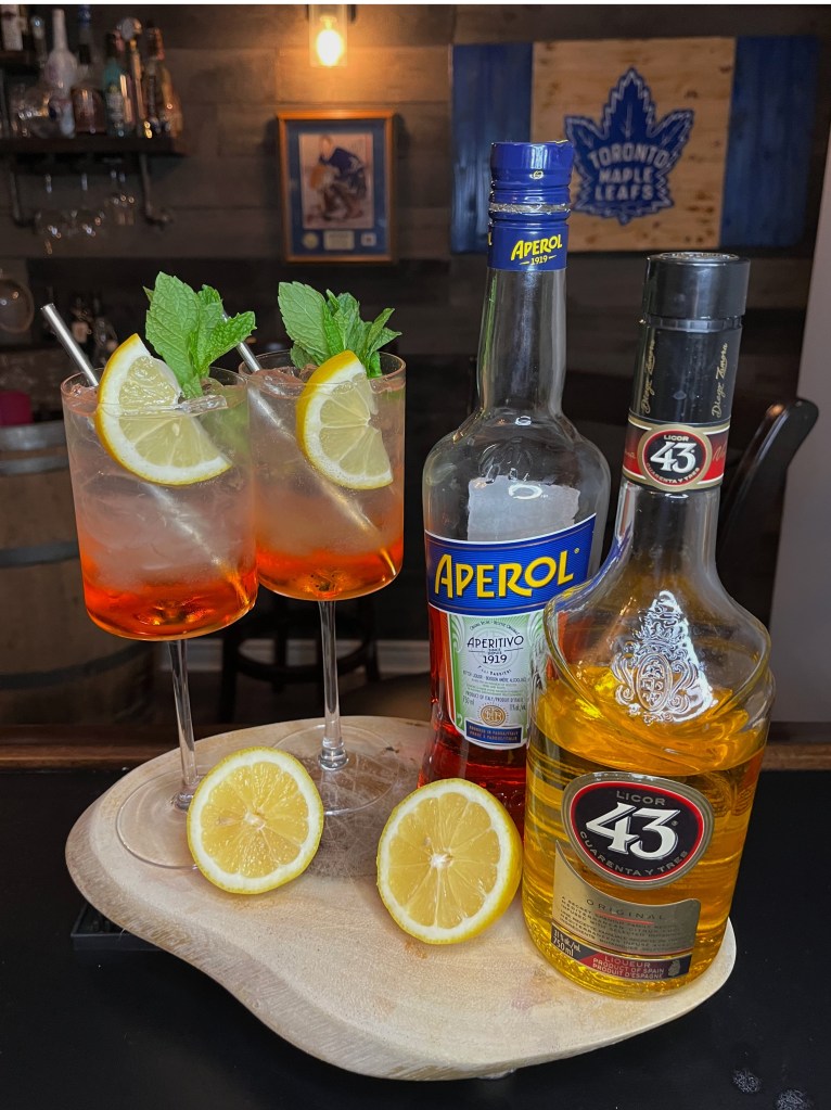 Bright, bubbly highball-ish cocktail over ice featuring vibrant Aperol and golden Licor 43, zesty fresh lemon juice, smooth vodka, Sprite fizz, finished with a lemon wheel and aromatic mint sprig garnis

