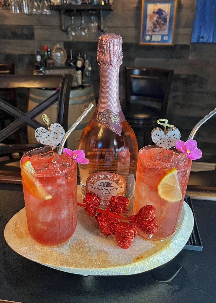Layered pink champagne cocktail in a tall Collins glass: vibrant Chambord liqueur base, fresh lemon juice and simple syrup, topped with bubbly pink champagne and a striking float of Empress Elderflower Rose Gin. Garnished with a dragon fruit heart slice, fresh lemon wheel, and edible flower for a romantic, elegant presentation

