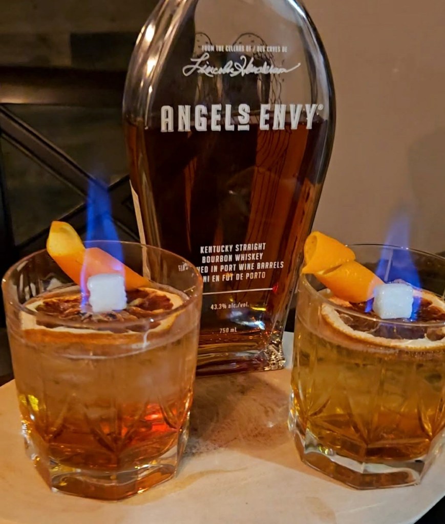 A rocks glass filled with an amber bourbon cocktail topped with fizzy club soda, garnished with an expressed orange peel, featuring a dramatic blue flame burning atop a sugar cube soaked in absinthe and placed on a dried orange wheel perched on the rim.

