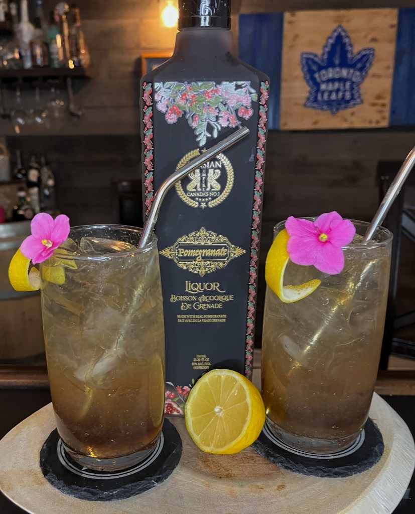 Deep crimson gothic cocktail in a tall Collins glass filled with ice, topped with club soda fizz, garnished with a lemon twist and delicate violet flower—mysterious pomegranate whiskey romance in a glass.

