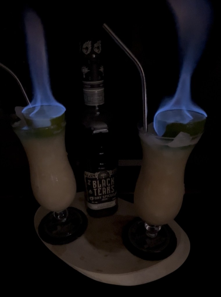 "Refreshing tiki-style cocktail in a hurricane glass filled with crushed ice, made with spiced rum, cognac, orgeat syrup, fresh lemon, lime, and grapefruit juices, double-strained for smoothness, topped with a dramatic flaming lime half garnish soaked in Chartreuse (or absinthe) over a sugar cube, creating a fiery presentation before the flame subsides

