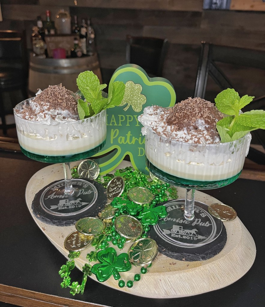 St. Patrick's Day chocolate Blarney Stone cocktail in a chilled coupette glass: a large ice ball coated in chocolate shell, topped with whipped cream, grated chocolate, and a fresh mint sprig garnish. The drink features layers of green creme de menthe, chocolate liqueur, Baileys Irish Cream, and cream for a creamy mint-chocolate sip. Guests kiss the chocolate 'Blarney Stone' for good luck before stirring and enjoying.

