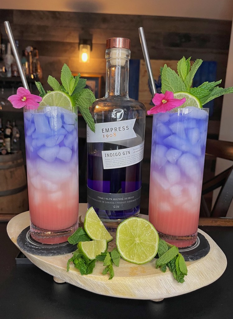 "Vibrant layered cocktail in a tall Collins glass: crushed ice base with pink-red Campari-orGeat-lime mix, topped with sparkling club soda and a striking purple Empress Gin float, garnished with lime wheel, fresh mint sprig, and delicate violet petal.

