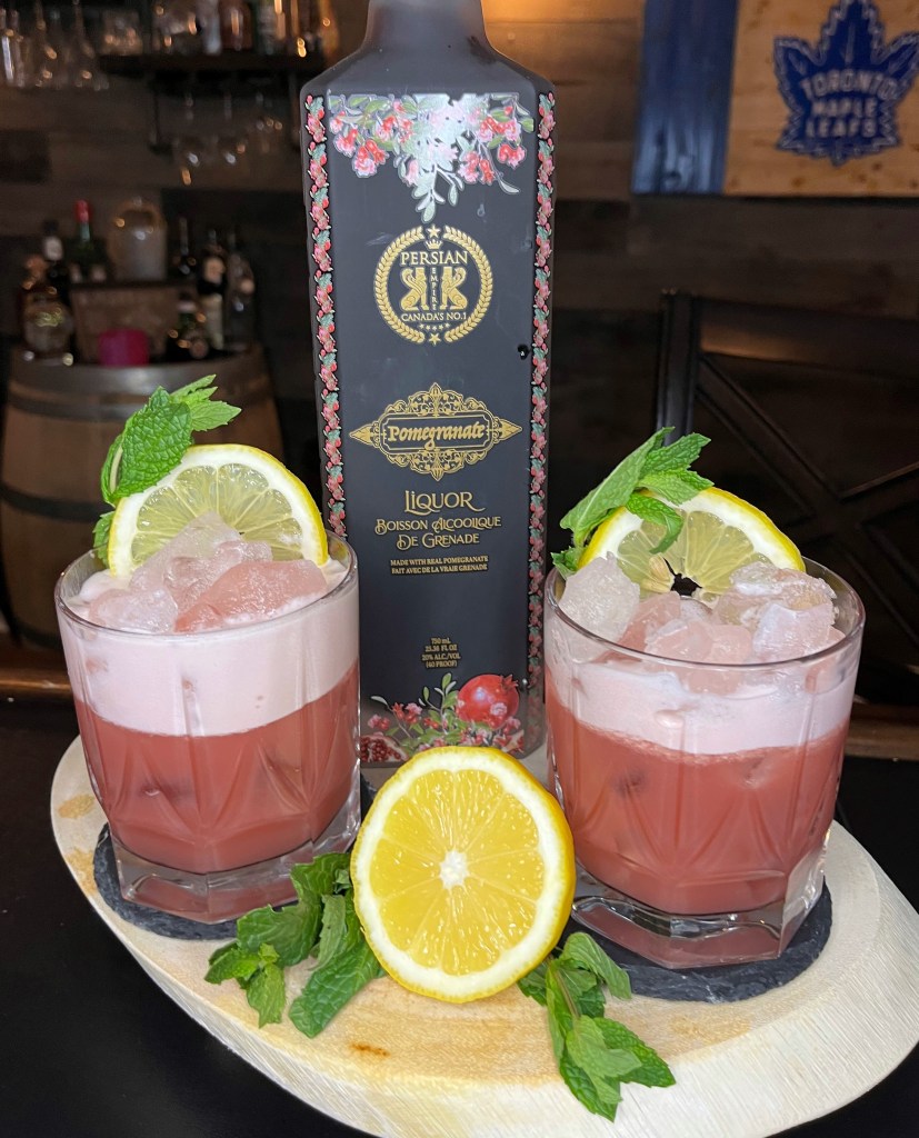 Beautifully balanced bourbon cocktail in a 12oz tumbler with vibrant pink hue from pomegranate, topped with silky egg white foam, garnished with a fresh lemon wheel and mint sprig
