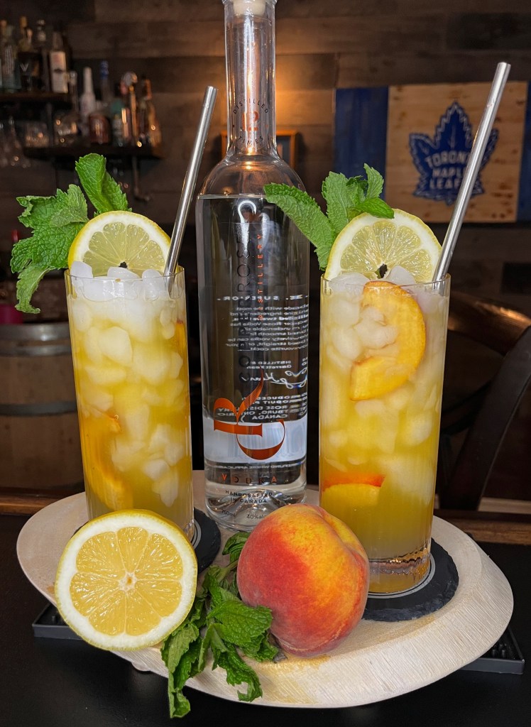 "A refreshing 'The Hammer' cocktail in a tall Collins glass filled with crushed ice, fresh peach slices, topped with club soda, garnished with a lemon wheel and mint sprig. Made with Copper Rose Vodka, lemonade, and peach nectar at Miller's Amish Pub.

