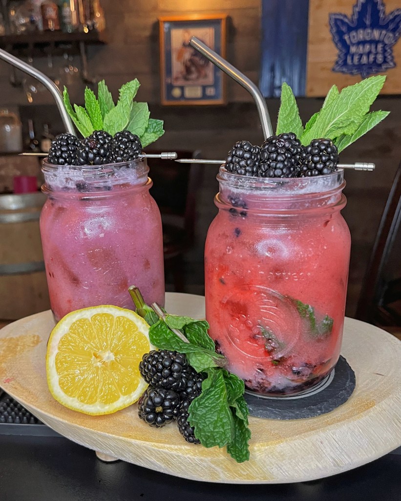 "Blackberry Coconut Mojito served in a mason jar glass, featuring muddled fresh blackberries and mint, creamy coconut, coconut rum, fresh lemon juice, and club soda. Garnished with a whole blackberry and mint sprig.

