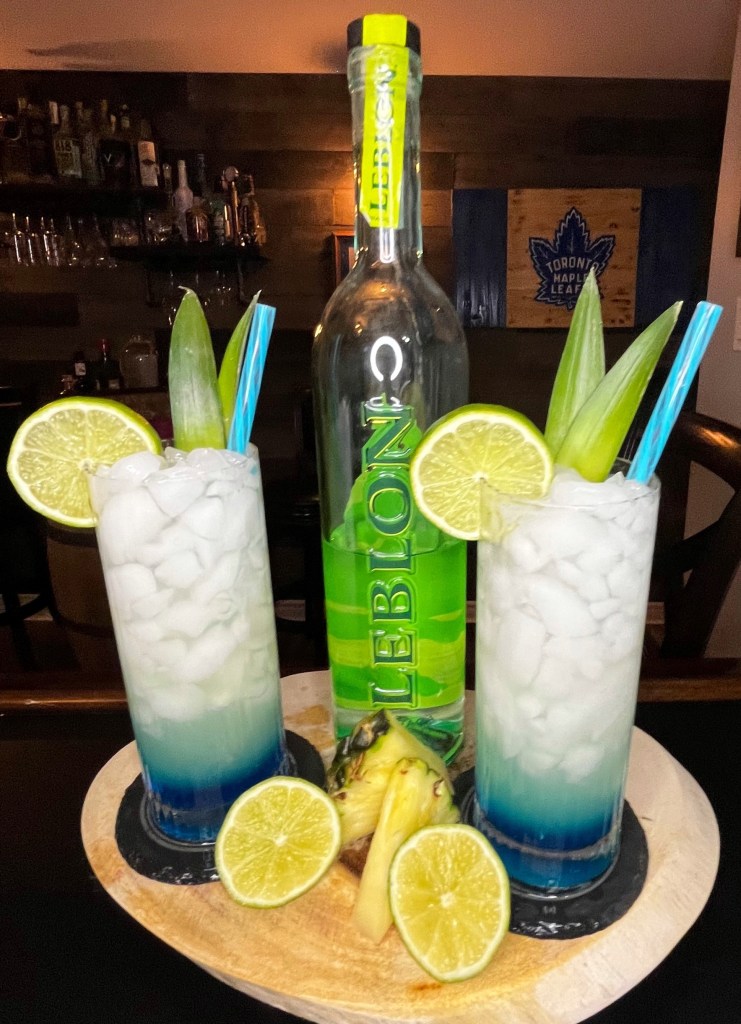 A vibrant tropical cocktail in a 14oz Collins glass, layered with blue curaçao at the bottom and topped with sparkling club soda. Made with cachaça, fresh lime juice, ginger liqueur, and pineapple juice, shaken and strained over crushed ice. Garnished with a fresh lime wheel and pineapple fronds.

