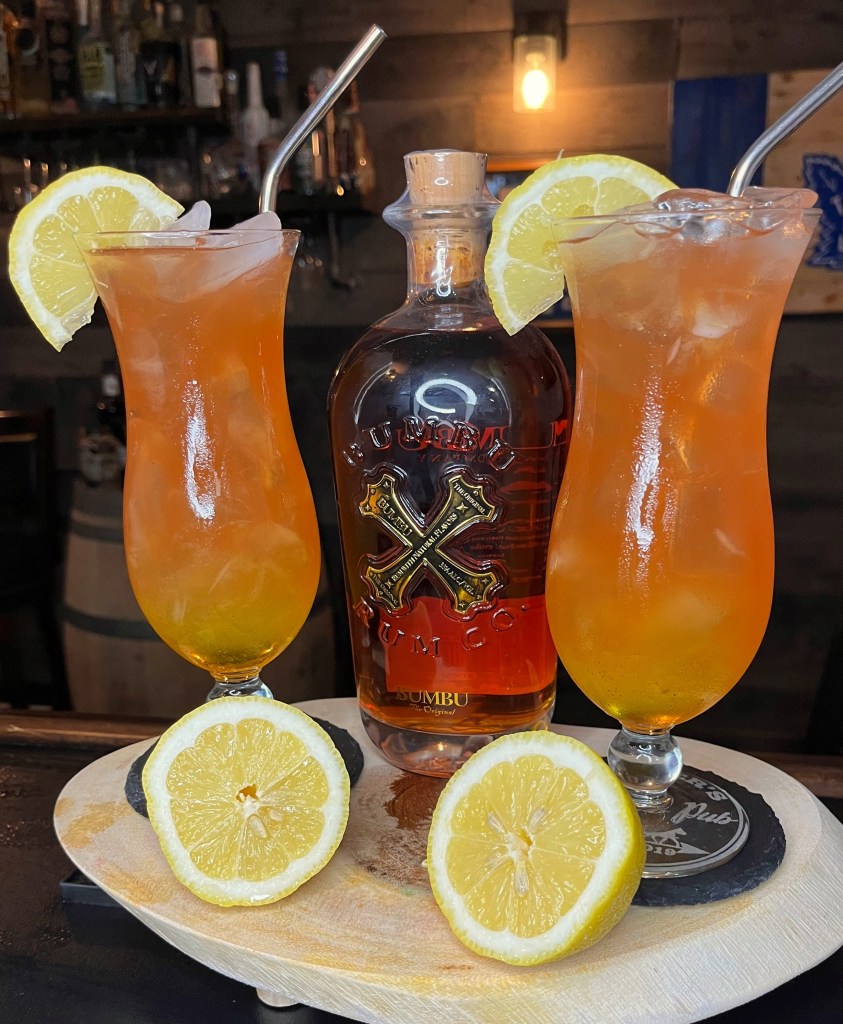 A deep, complex, and refreshing rum cocktail with subtle tropical notes and zero cloying sweetness.  Made with spiced rum, banana liqueur, fresh lemon juice, coconut water, club soda, cherry syrup, and a hint of cardamom. Served over ice in a hurricane glass and garnished with fresh lemon.  Perfect for sipping in the sun!  

