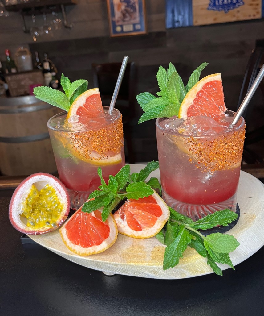 Millers Amish Pub's Passionate Paloma cocktail in a Tajin-rimmed tumbler, featuring tequila, fresh passionfruit, grapefruit, blackberry syrup, and club soda, garnished with a grapefruit wheel and mint sprig."