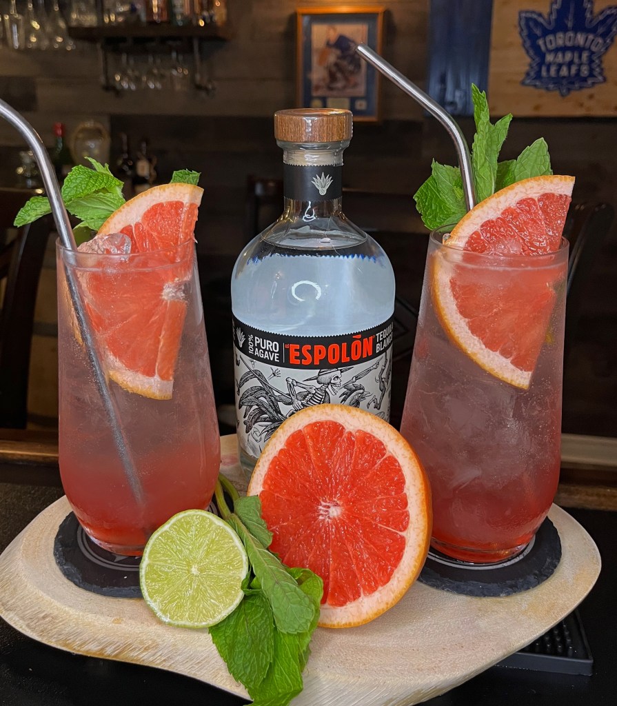Refreshing Paloma-style cocktail served in a tall Collins glass filled with ice. The vibrant pink drink is made with tequila, Campari, fresh grapefruit and lime juice, simple syrup, and topped with club soda. Garnished with a salted grapefruit half-wheel and fresh mint sprig

