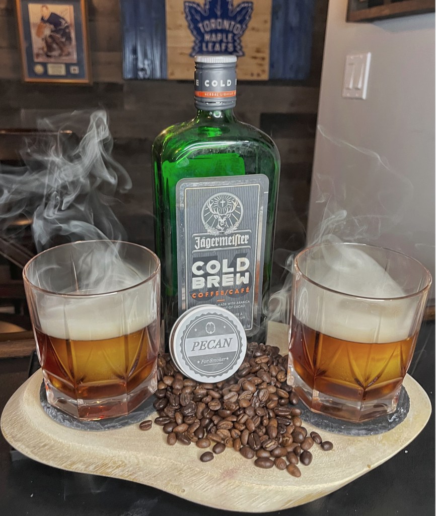 Smoky Espresso Martini in a rocks glass with a large crystal-clear ice sphere. The cocktail features vodka, fresh espresso, Jägermeister Coffee Liquor, Frangelico hazelnut liqueur, and simple syrup, dramatically smoked with coffee beans and pecan wood.

