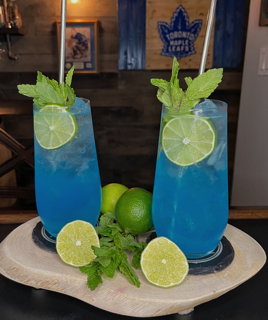 Vibrant electric-blue cocktail in a Collins glass filled with ice, topped with a refreshing Sprite fizz, and garnished with fresh mint and a lime wheel. Bright, tropical Blue Patio Bliss cocktail on a sunny patio setting.

