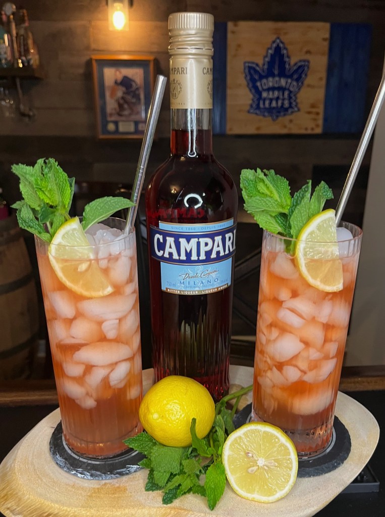 The Secret Agent Man cocktail in a tall Collins glass filled with crushed ice, featuring a bold reddish-amber mix of whiskey, Campari, and sweet vermouth, topped with spicy ginger beer and garnished with a fresh lemon wheel and vibrant mint sprig

