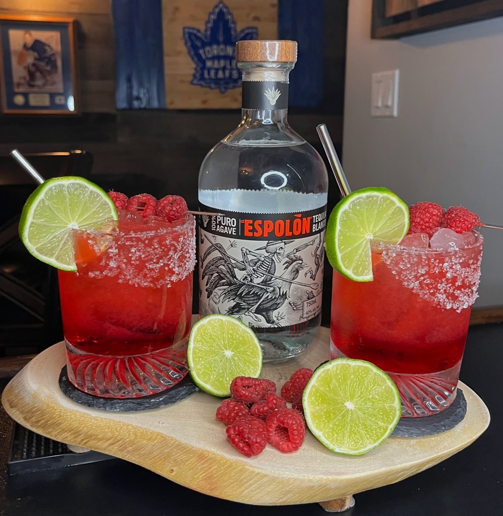 A vibrant pink and red Sour Raspberry Margarita cocktail in a classic margarita glass, garnished with fresh raspberries and lime, served at Miller's Amish Pub. Recipe #447 – a fun, fruity twist on the traditional margarita for Cinco de Mayo.

