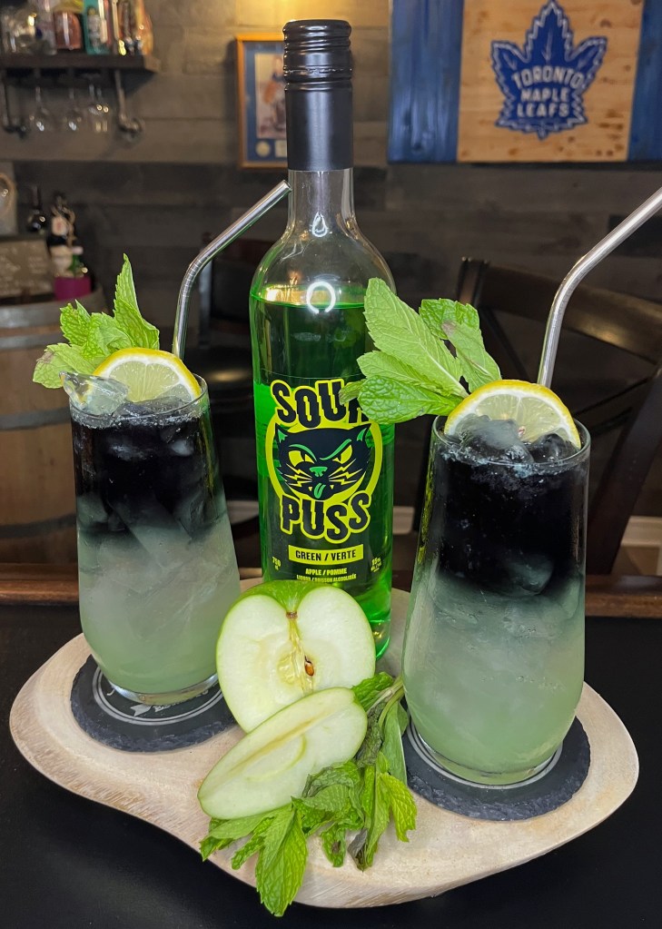 reen Apple Eclipse cocktail in a tall glass: vibrant green base made with vodka, Green Apple Sourpuss, and fresh muddled green apple, topped with a dramatic black vodka float creating a beautiful dark-to-emerald gradient. Garnished with apple slice and mint