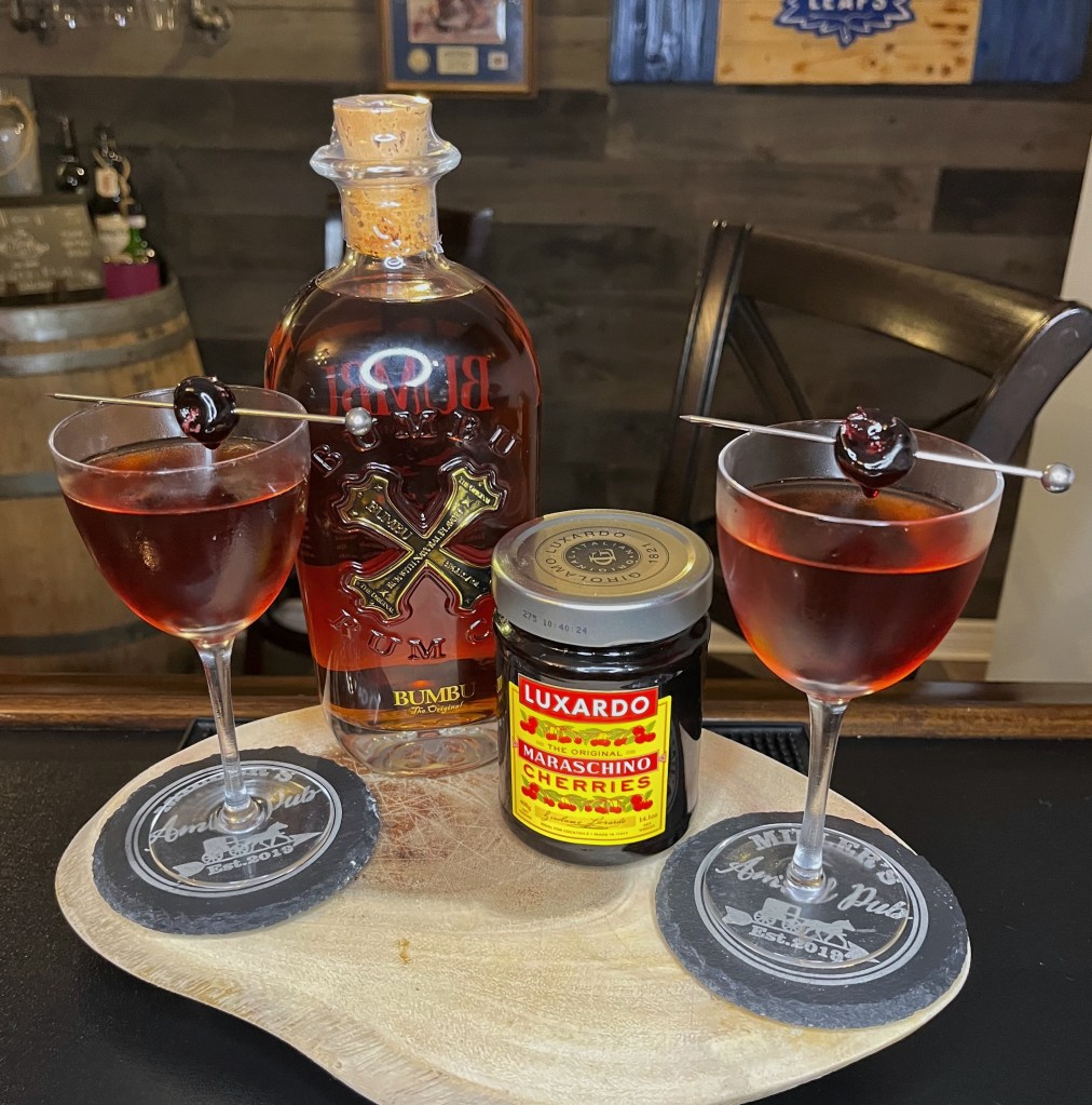 A refined, deep amber rum cocktail served in a chilled Nick & Nora glass and garnished with a glossy cocktail cherry. Made with Bumbu Rum, sweet vermouth, Luxardo cherry liqueur, cherry brandy, and Angostura bitters.

