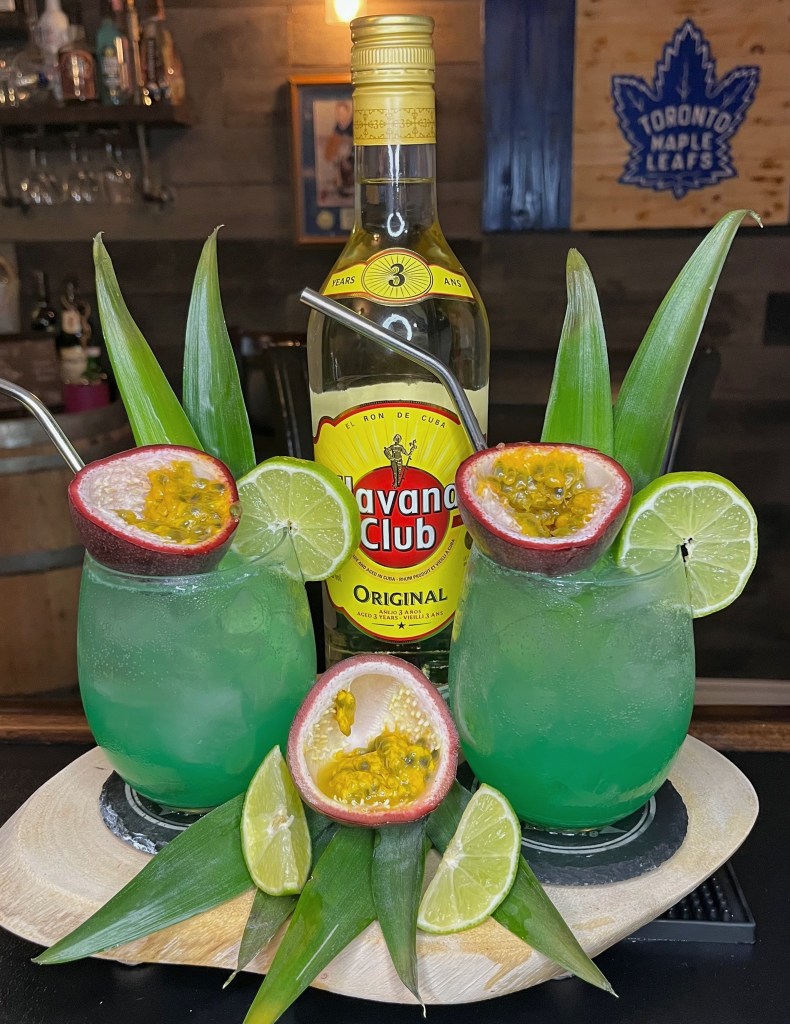 Paradise Patio Passion cocktail at Millers Amish Pub – a vibrant blue and orange rum cocktail served in a large stemless wine glass, topped with Sprite and garnished with pineapple fronds, half a passion fruit, and a fresh lime wheel

