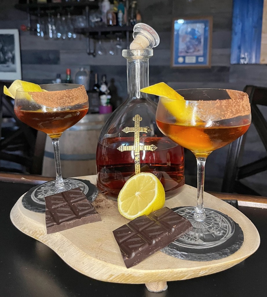 Velvet Glove cocktail in a chilled coupe glass. A rich amber-colored classic made with cognac, Lillet, and crème de cacao. Features a partial cocoa rim and a long elegant lemon twist garnish