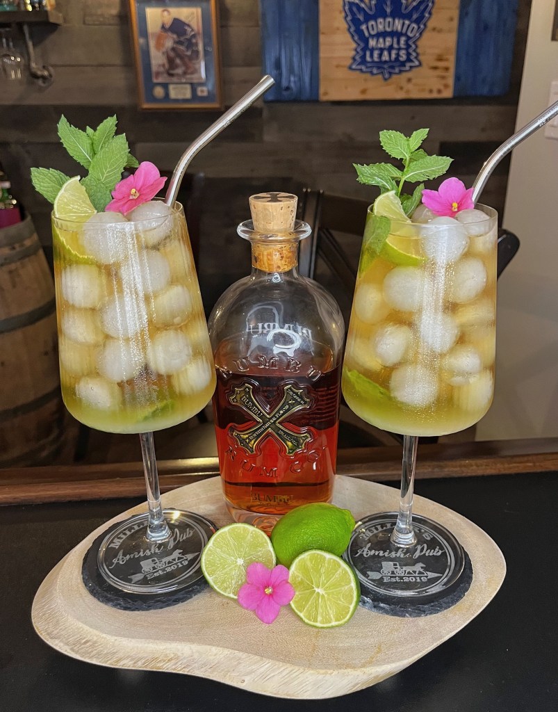 Amish Mojito in a stemmed glass: spiced rum, ginger ale, fresh lime, and muddled mint over ice, garnished with mint sprig, lime wedge, and violet petal. Miller’s Amish Pub Cocktail Recipe No. 465 – a summer favorite.