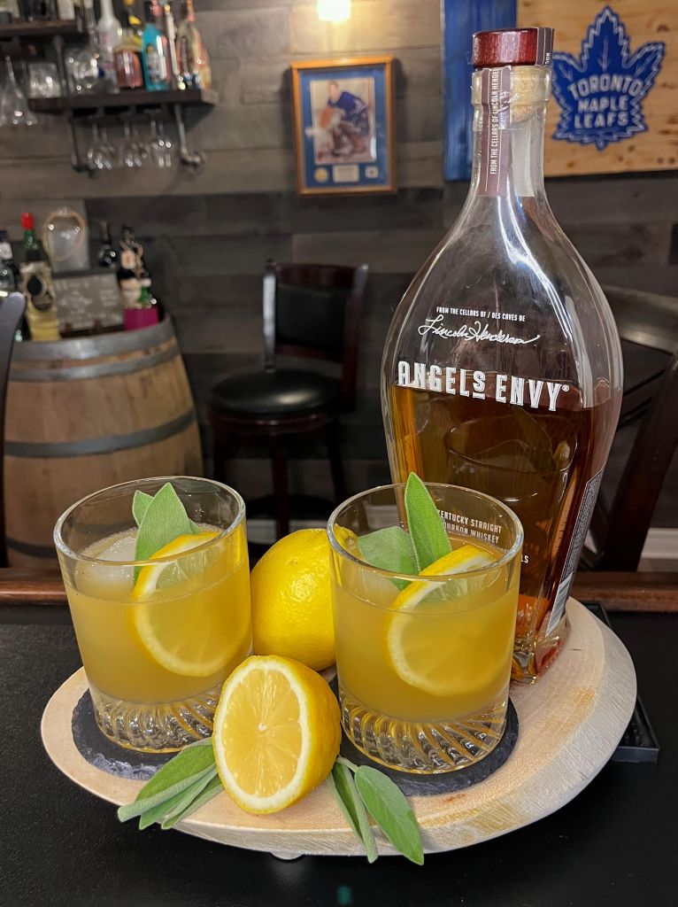 Two yellow bourbon cocktails over ice in rocks glasses, garnished with fresh sage and a lemon wheel next to a bottle of Angel's Envy bourbon and fresh lemons on a serving tray.