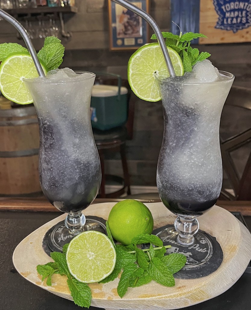 Two hurricane glasses filled with a Frozen Transfusion cocktail slushie, showing a dark purple grape juice layers swirling with white blended ice. The glasses are garnished with fresh lime wheels, mint sprigs, and curved metal straws, sitting on a wooden serving tray with fresh lime halves and mint leaves in a home bar setting.