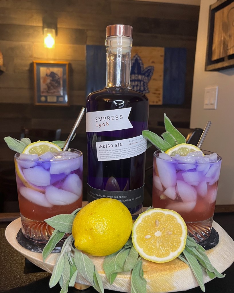 Two glasses of the Garden Zephyr cocktail over ice, showing a purple Empress Gin float layered over a pink juice base. The drinks are garnished with lemon and sage on a wooden tray next to an Empress Gin bottle and fresh lemons against a bar background.
