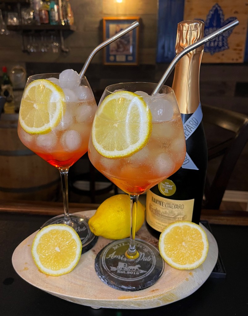 Two large stemmed wine glasses filled with a vibrant orange and pale yellow layered spritz cocktail over round ice cubes. The drinks are garnished with fresh lemon wheels and curved metal straws, arranged on a wooden tray with fresh lemon halves and a bottle of Ravine Vineyard sparkling wine against a rustic pub background.
