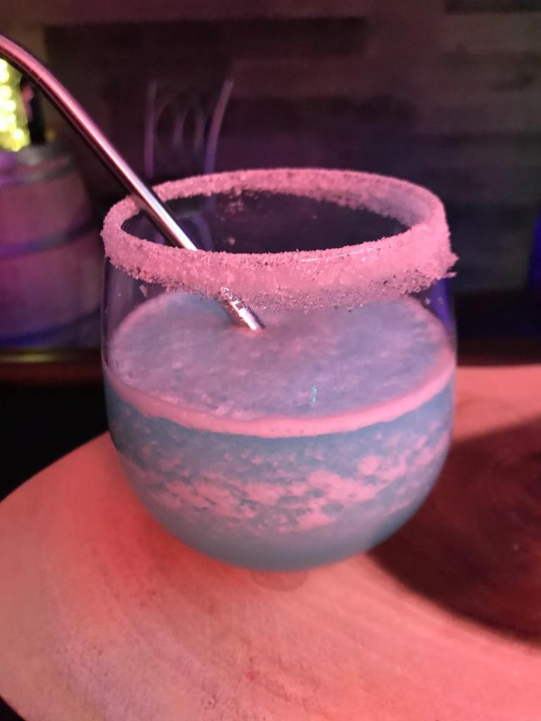 A bright blue frozen blended cocktail slushie served in a sugar-rimmed stemless wine glass and topped with shaved coconut flakes.
