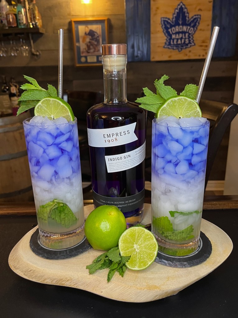 Two layered gin and lime cocktails with crushed ice, fresh mint leaves, and a purple gin float next to a bottle of Empress 1908 Gin and a fresh lime on a wooden tray