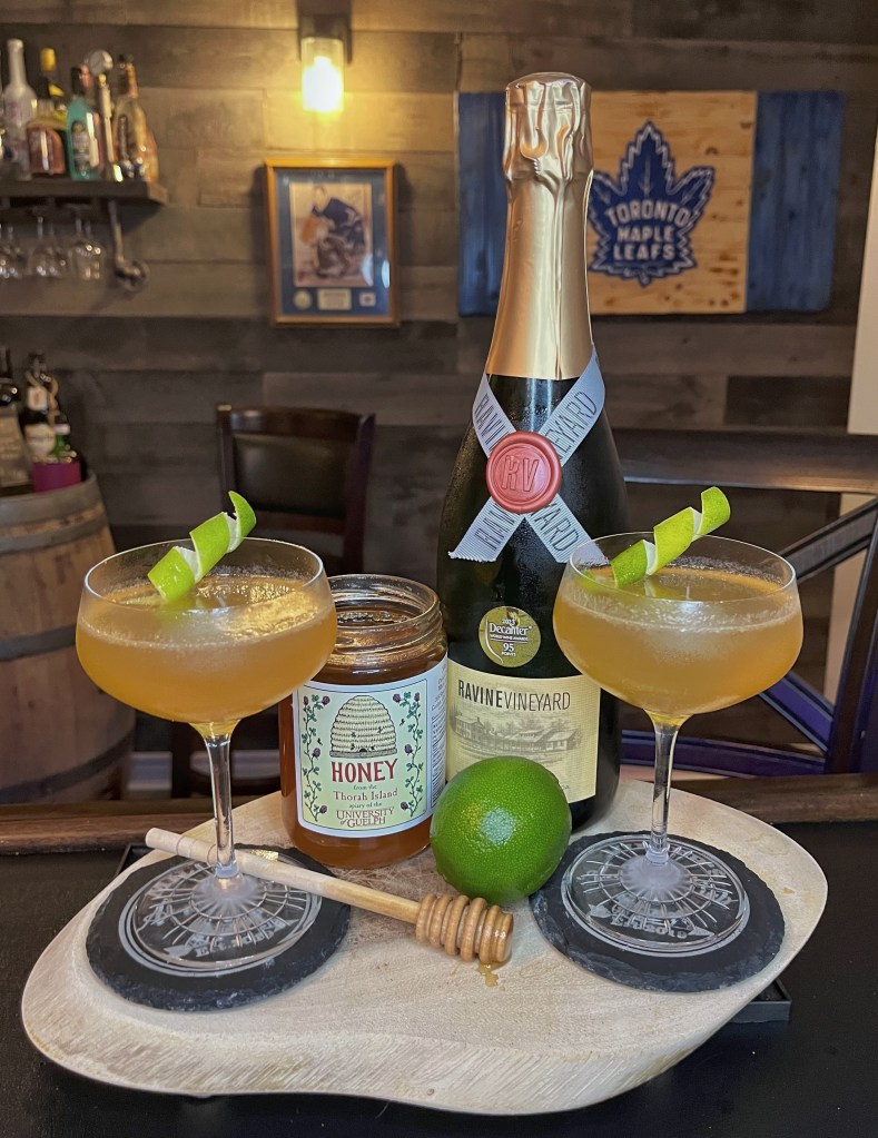Close-up of two amber-colored Air Mail cocktails in coupe glasses garnished with lime twists, arranged on a wooden platter alongside a jar of Pelee Island wildflower honey, a physical wooden honey dipper, and a bottle of Ravine Vineyard sparkling wine