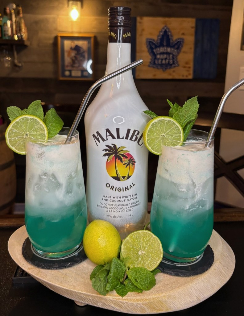Two teal Malibu rum cocktails in Collins glasses topped with white dark rum sea foam floats, garnished with lime wheels and fresh mint.