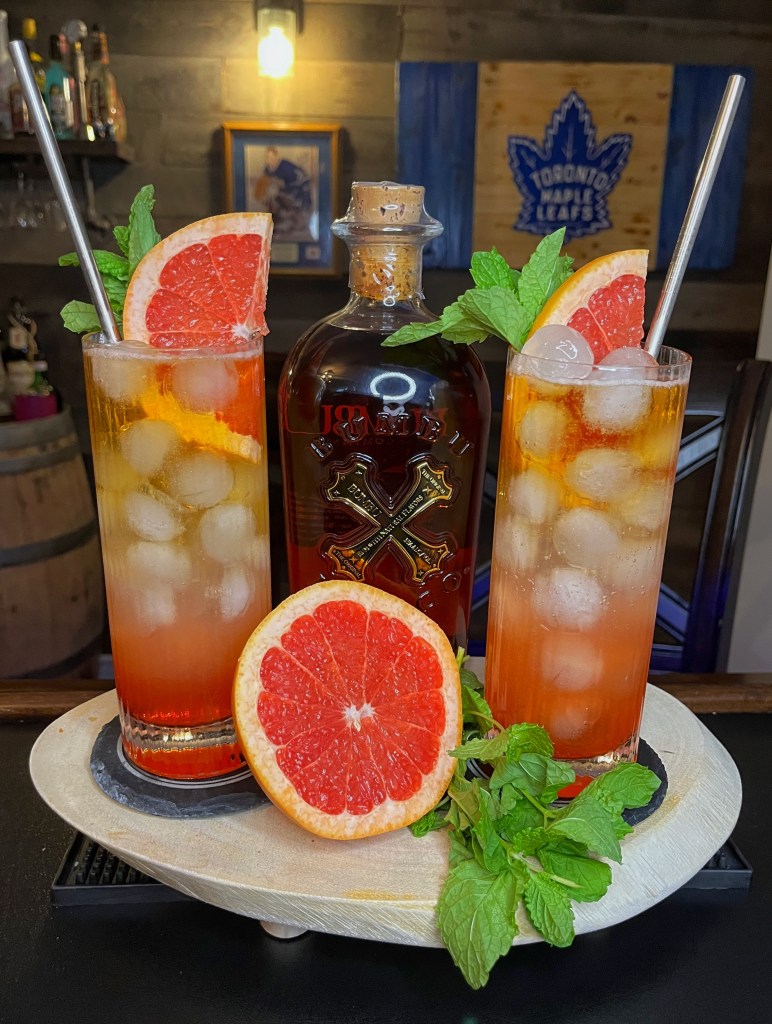 Two tall highball glasses filled with a layered rum and grapefruit cocktail over round ice cubes, featuring a crimson base gradient fading into amber. The drinks are garnished with fresh ruby grapefruit wedges, mint sprigs, and metal straws on a wooden tray next to a bottle of Bumbu Rum, a cut grapefruit half, and fresh mint against a home bar background.