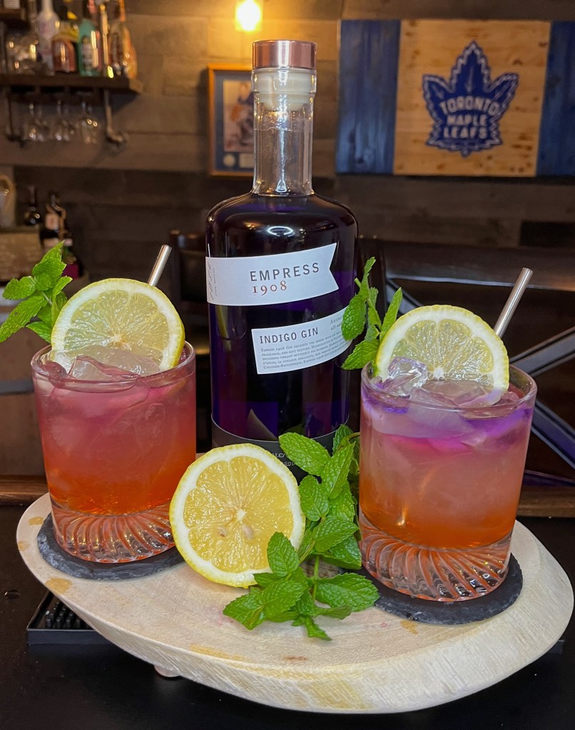 Two sunset-layered Flirty Little Whisper gin cocktails in rocks glasses garnished with lemon wheels and mint sprigs next to a bottle of Empress 1908 Indigo Gin.