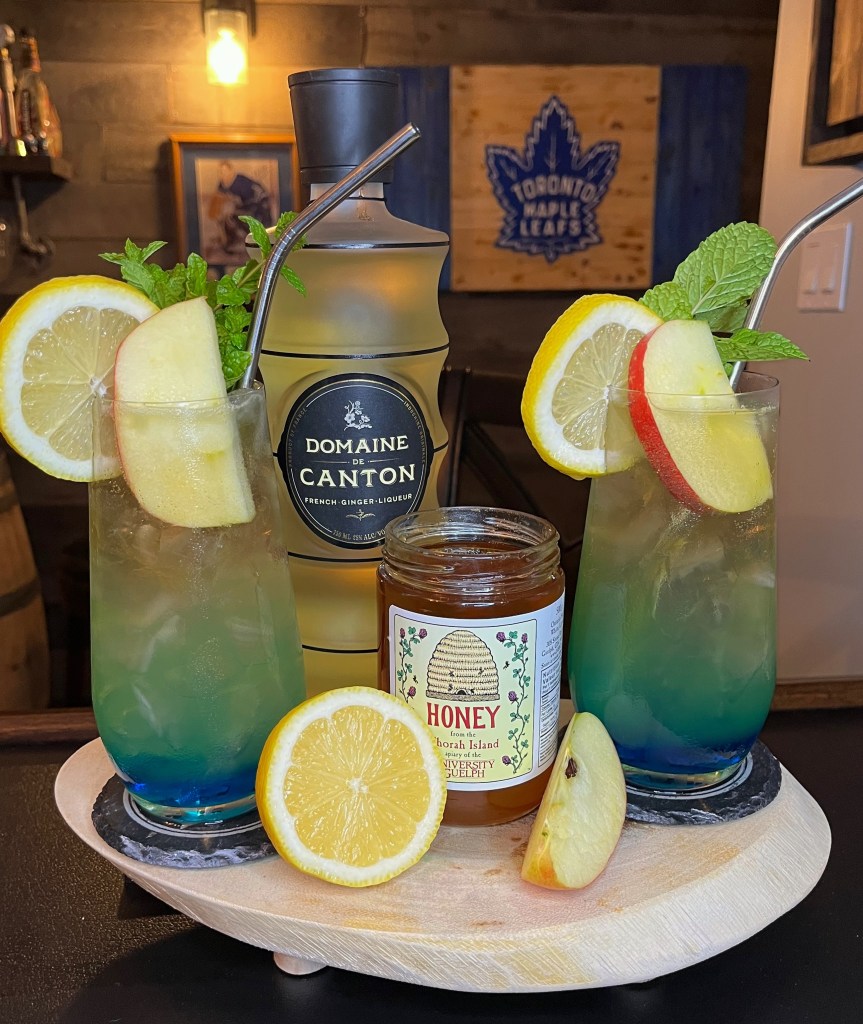 Two tall glasses filled with a layered Ginger Honey Crisp cocktail over ice, featuring a bright blue base fading into a golden-green juice blend. The drinks are garnished with fresh apple slices, lemon wheels, and mint sprigs, styled on a wooden tray with a jar of honey, a bottle of Domaine de Canton, and fresh fruit.