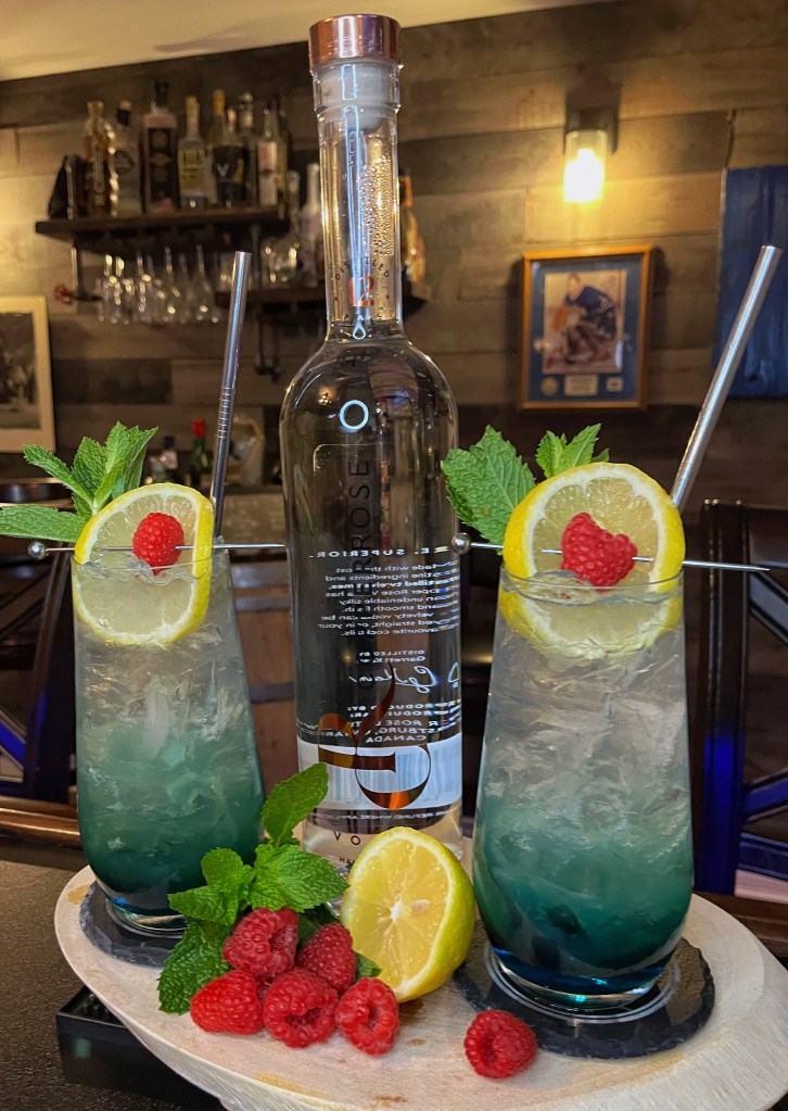 Two vibrant teal-layered Jungle Fling cocktails served in Collins glasses with lemon wheels, fresh raspberries, and mint sprigs next to a bottle of Copper Rose Vodka.