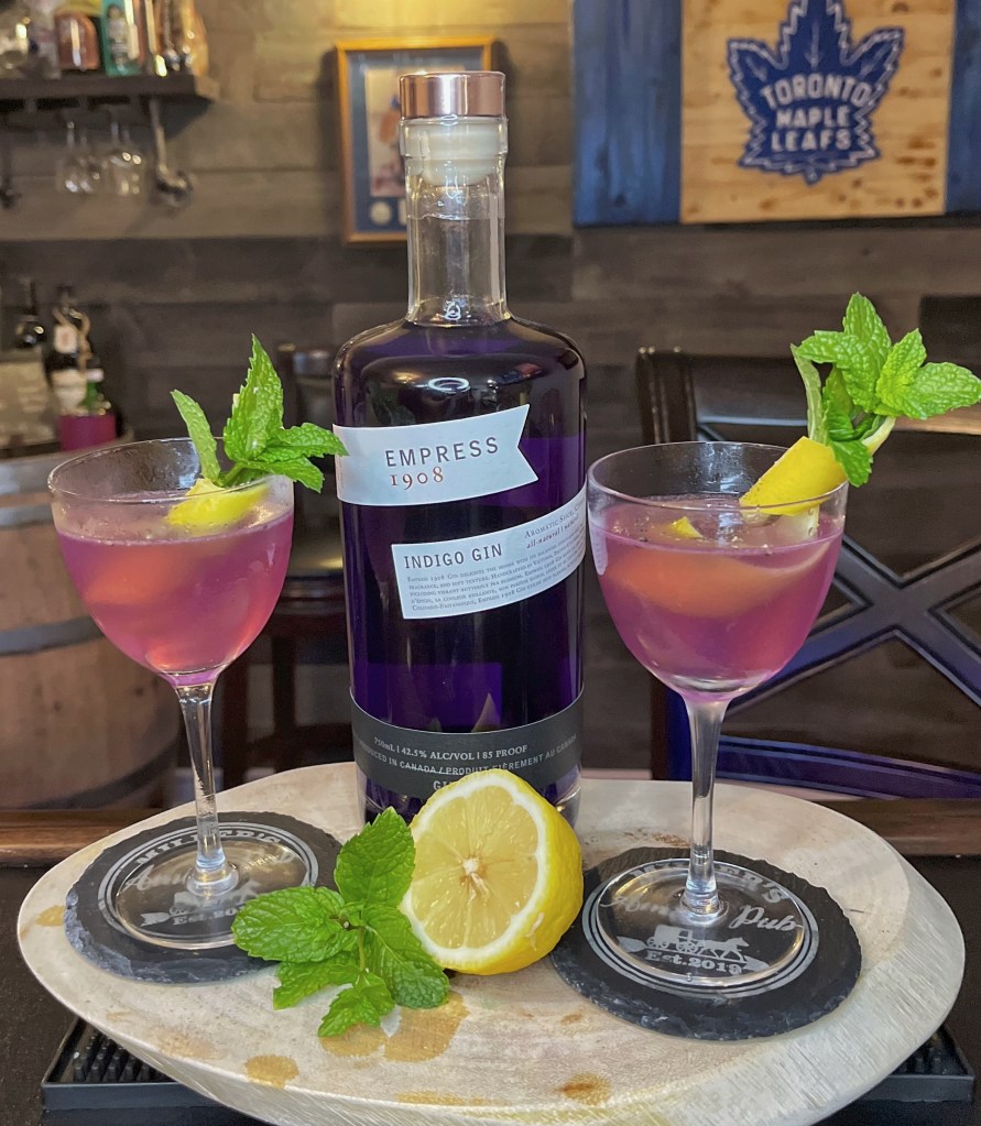 A bottle of purple Empress 1908 Indigo Gin sits between two elegant coupe glasses filled with a vibrant lavender-pink cocktail. Each glass is resting on a black slate coaster and garnished with a fresh green mint sprig and a lemon peel twist. In the foreground, a halved fresh yellow lemon and loose mint leaves rest on a rustic wooden serving board.