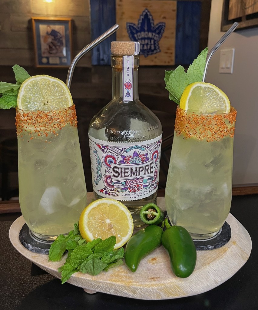 Two tall glasses of the Spicy Mexican Water Hazard cocktail over ice with a chili-salt rim. The drinks are garnished with fresh lime wheels and mint sprigs on a wooden tray next to a Siempre Tequila bottle, whole jalapeños, and fresh lemons against a pub background.