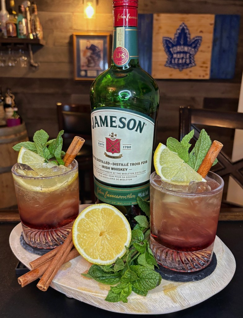 Two Autumn in the Éire fall whiskey cocktails in tumblers next to a Jameson Irish Whiskey bottle on a wooden bar tray. The layered red and golden drinks are garnished with a fresh lemon wheel, a cinnamon stick, and mint sprigs, surrounded by fresh cocktail ingredients.