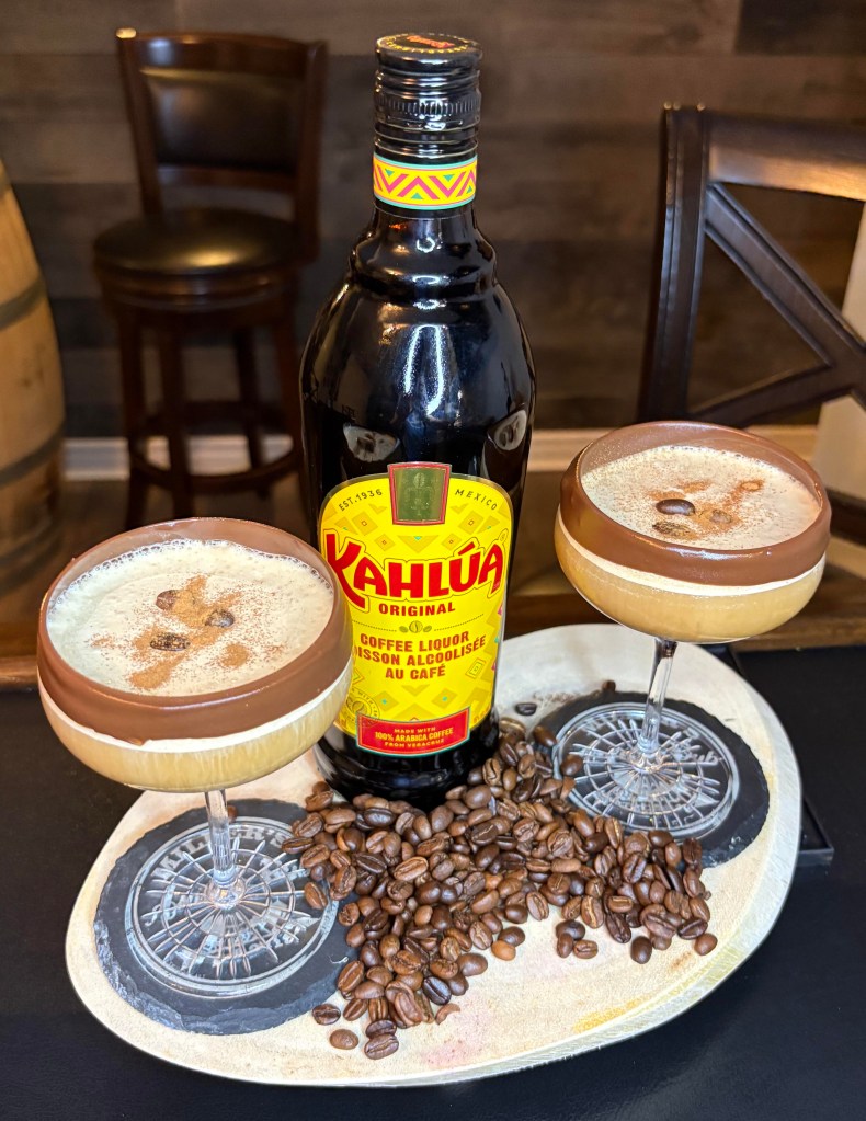 A chocolate cinnamon espresso martini in a coupe glass with a chocolate-dipped rim and coffee beans, next to a bottle of Kahlúa.