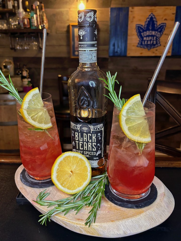 The Cozy Hoodie cocktail, a vibrant spiced rum drink garnished with fresh lemon and rosemary, served next to a bottle of Black Tears Dry Spiced Rum.