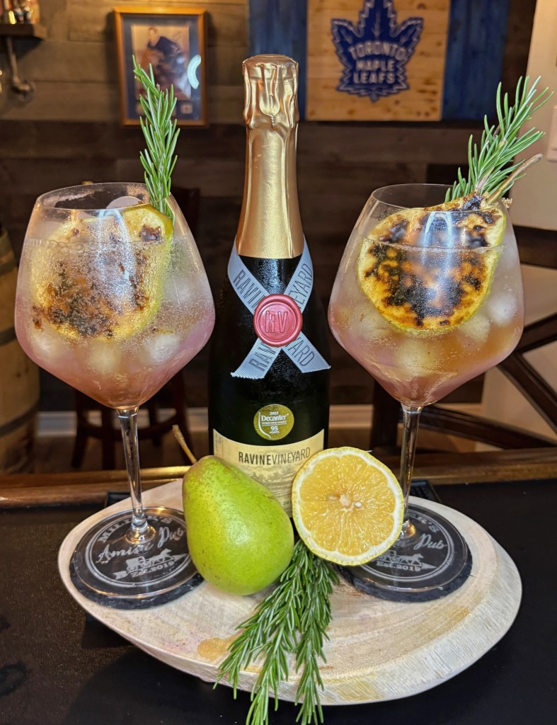 Sparkling pear and Empress Gin autumn cocktail in large wine glasses, garnished with a bruleed pear slice and rosemary, next to a bottle of Ravine Vineyard sparkling wine.