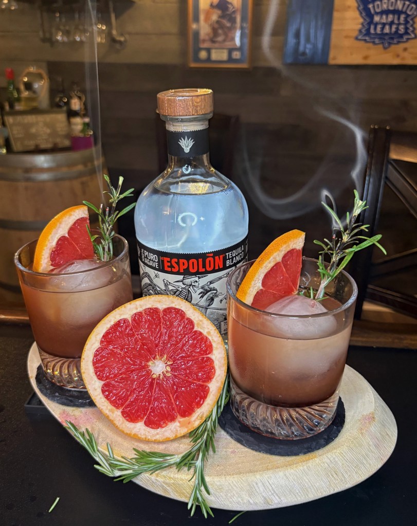 Two smoked grapefruit and rosemary tequila cocktails on a wooden board next to a bottle of Espolòn Blanco tequila, fresh grapefruit halves, and smoking rosemary sprigs.