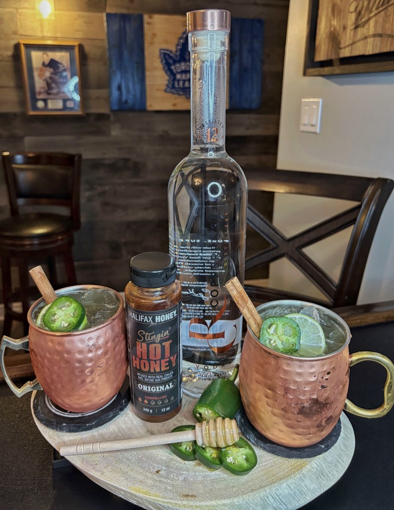 A Sweet Heat Mule cocktail served in a frosty copper mug on a rustic pub table. The apple cider and vodka cocktail is garnished with a green lime wheel, a fresh jalapeño slice, and a cinnamon stick, surrounded by fresh fall ingredients at Millers Amish Pub