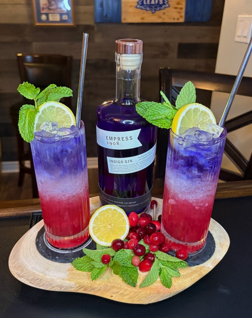A vibrant red, white, and blue layered cocktail in a Collins glass made with Empress 1908 Gin, cranberry juice, and club soda. The drink features a deep purple gin float on top, a clear club soda layer over crushed ice, and a ruby-red cranberry base. It is garnished with a fresh lemon wheel, a mint sprig, and scattered cranberries on a rustic wooden tray.